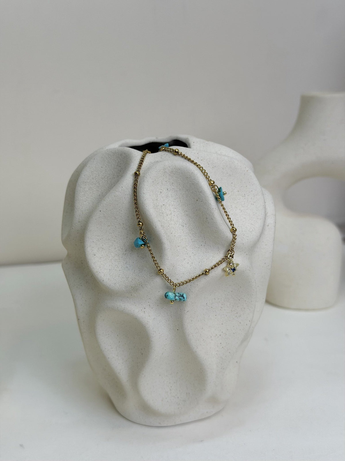 Paired Alvara Anklet in gold-tone, draped over a textured cream vase showing turquoise charms and a star