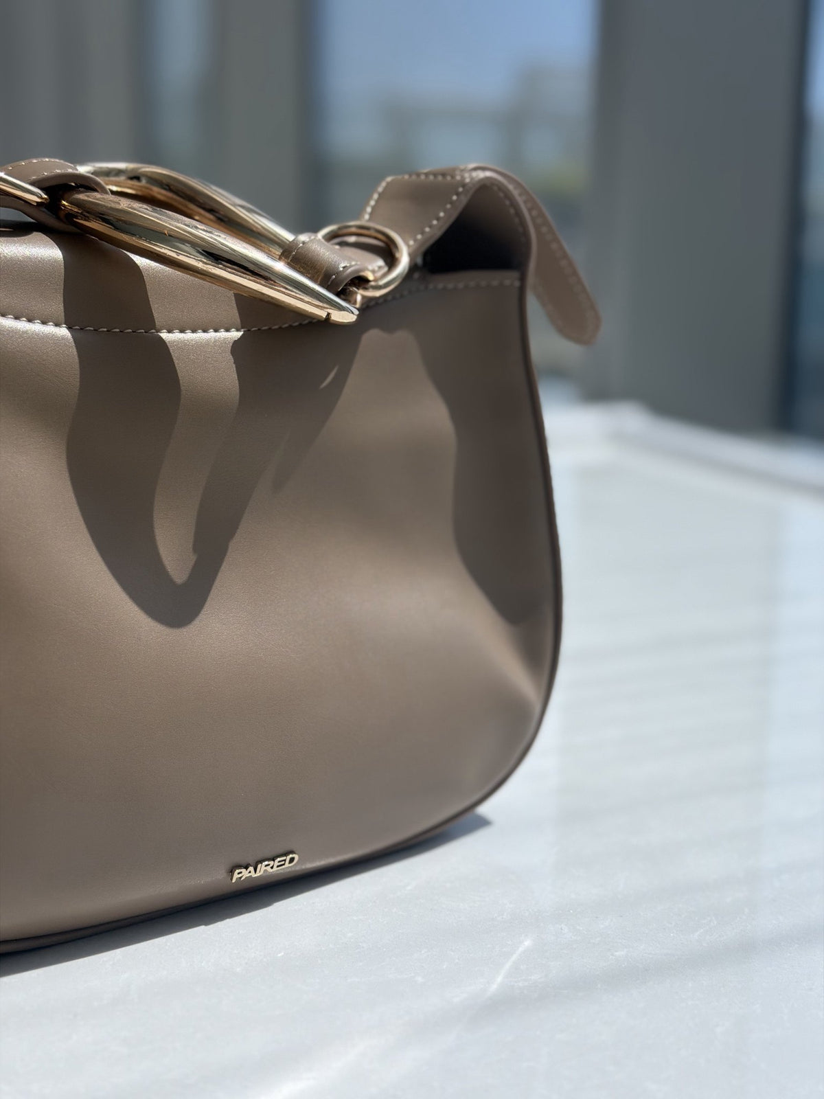 Paired Arden Soft Bag in taupe apricot, with a soft rounded shape and strap, ring handle at the top, on a white floor