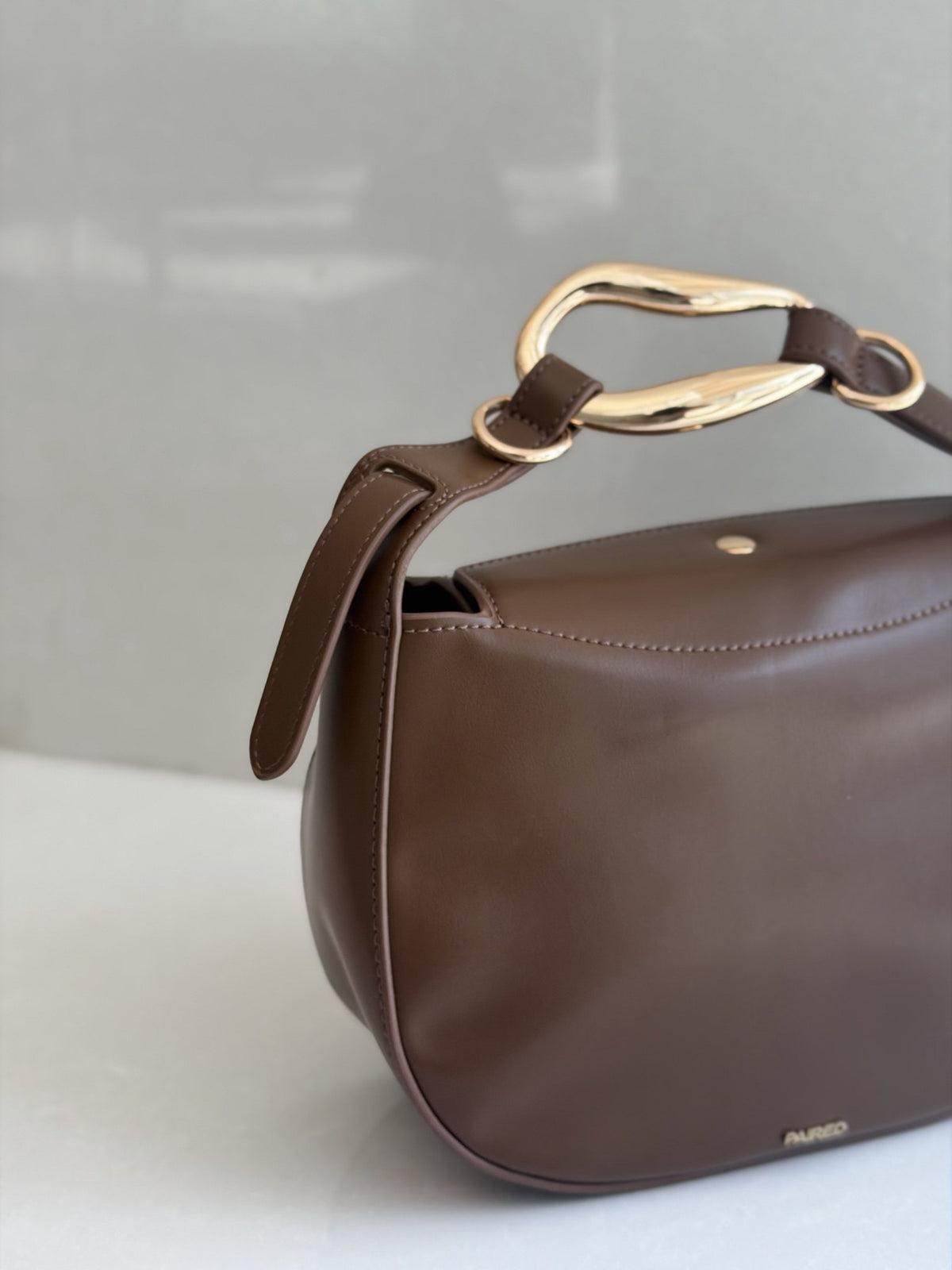 Paired Arden Soft Bag in coffee brown, seen from an angle with a sculptural gold-tone ring handle and small logo