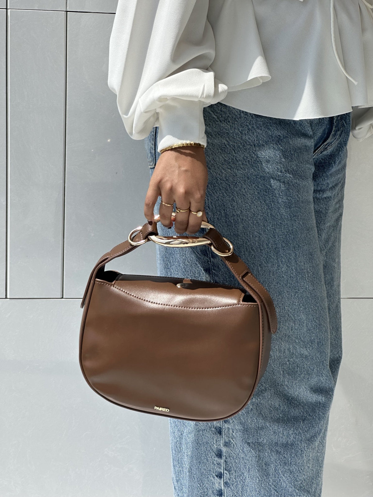 Paired Arden Soft Bag in coffee brown, held by the ring handle against jeans and a white blouse, photographed sideways