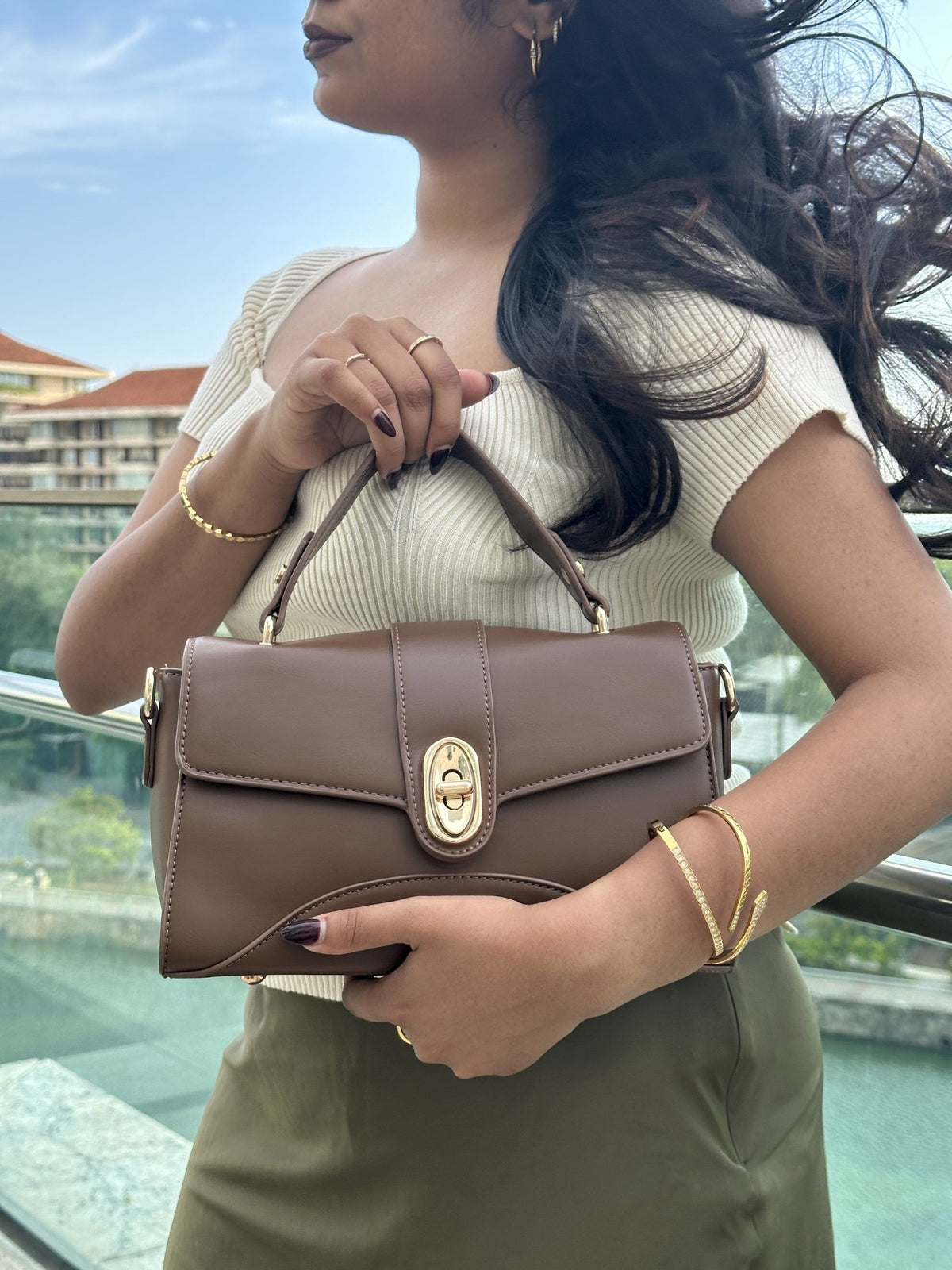 Paired Aurea Crest Bag in coffee brown, carried at the shoulder with a gold-tone turn-lock, photographed sideways
