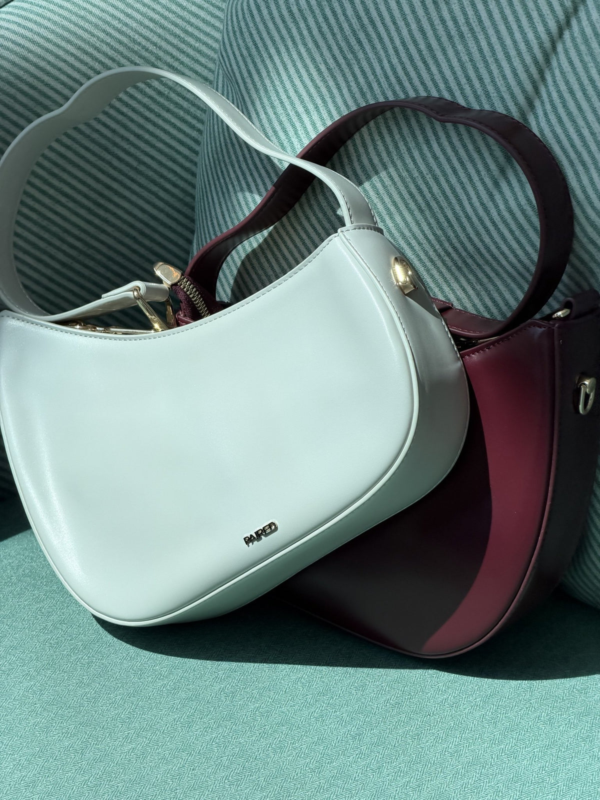Paired Clarissa Chain Bag in white and burgundy lying together on a teal armchair, with straps and chain links