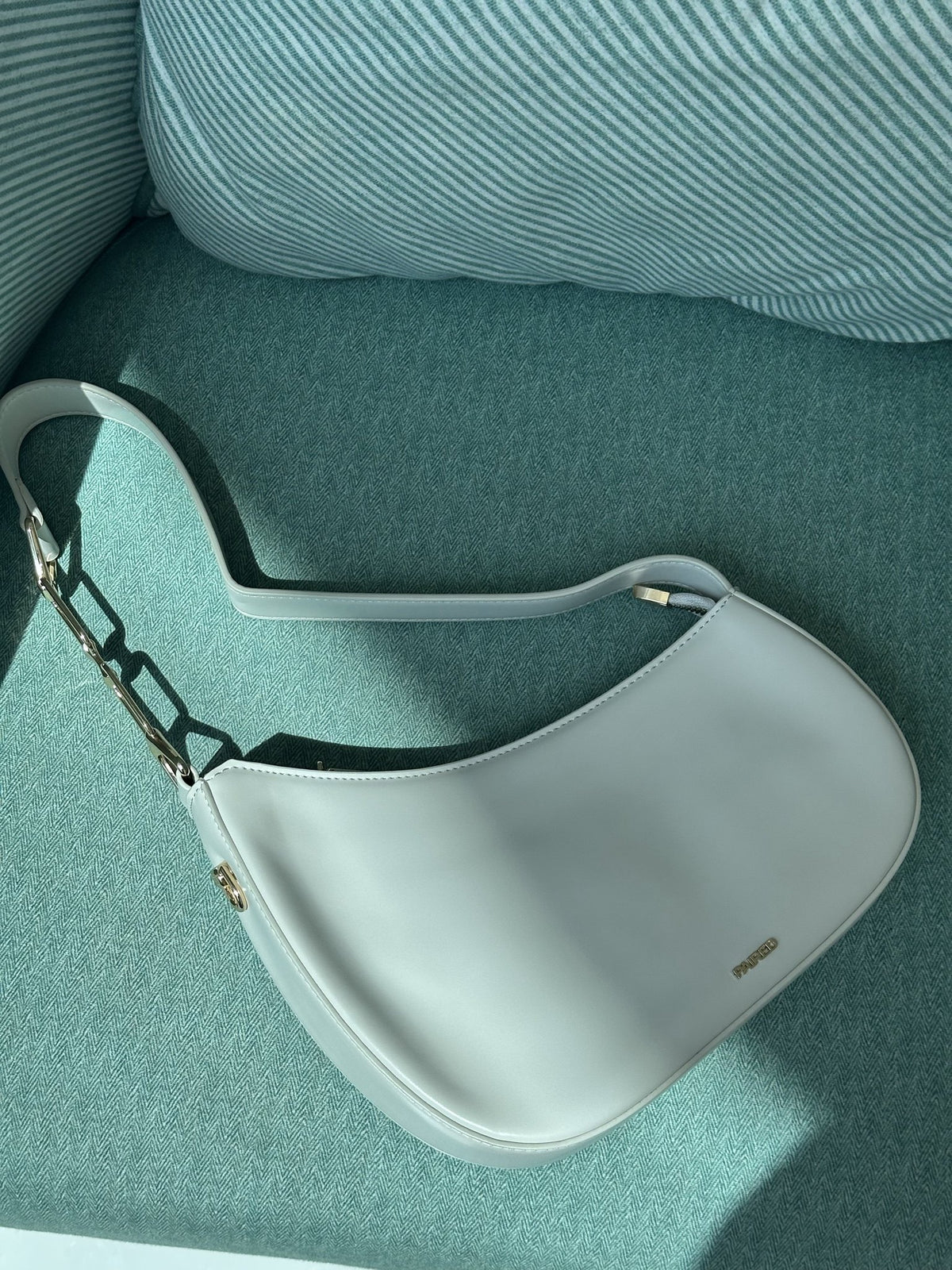 Paired Clarissa Chain Bag in white, a crescent shoulder bag with a flat strap and chunky chain link, lying on a teal armchair