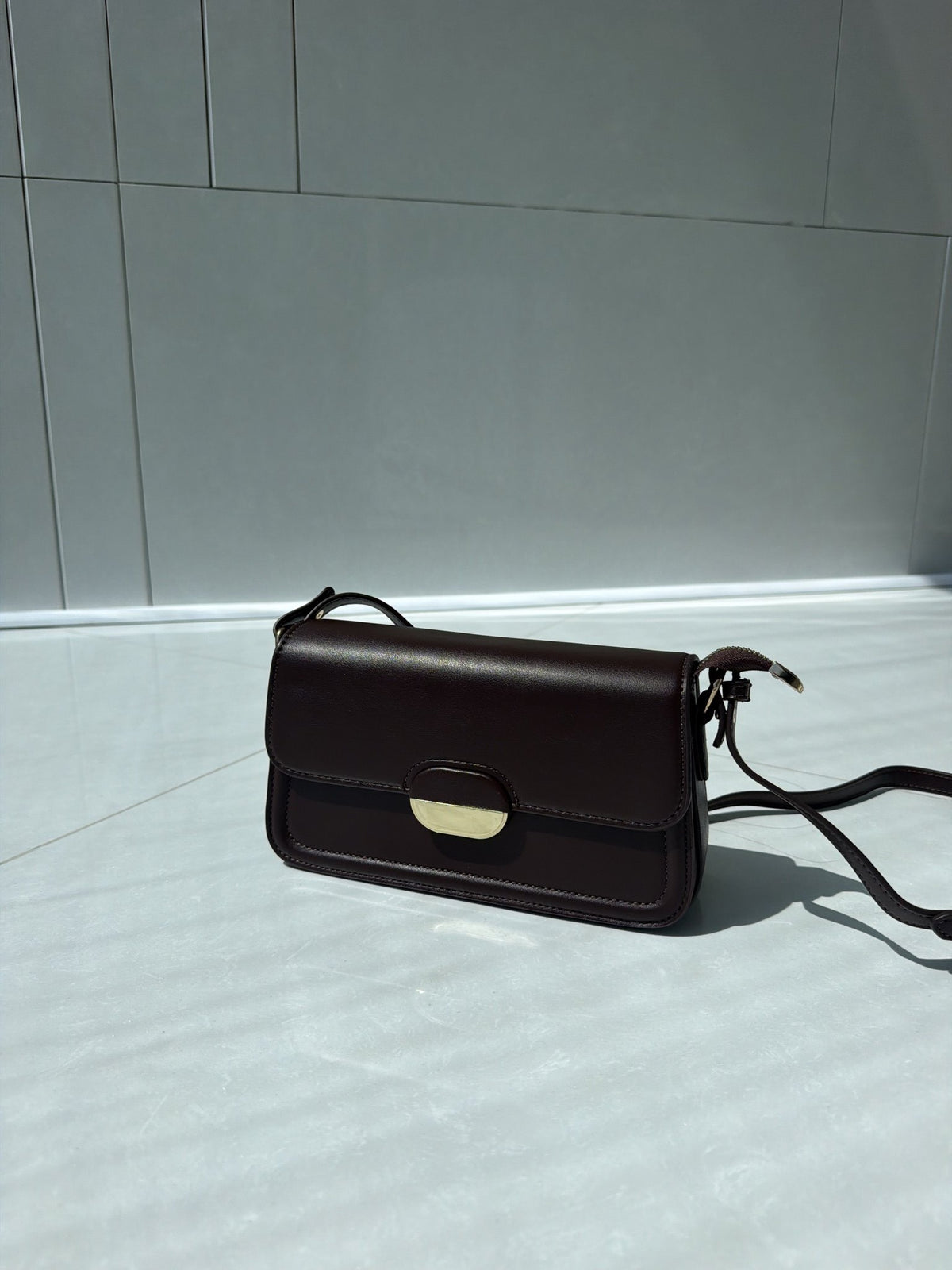 Paired Elise Lock Bag in coffee brown, hanging by its long strap against a pale wall, showing the flap and lock plate