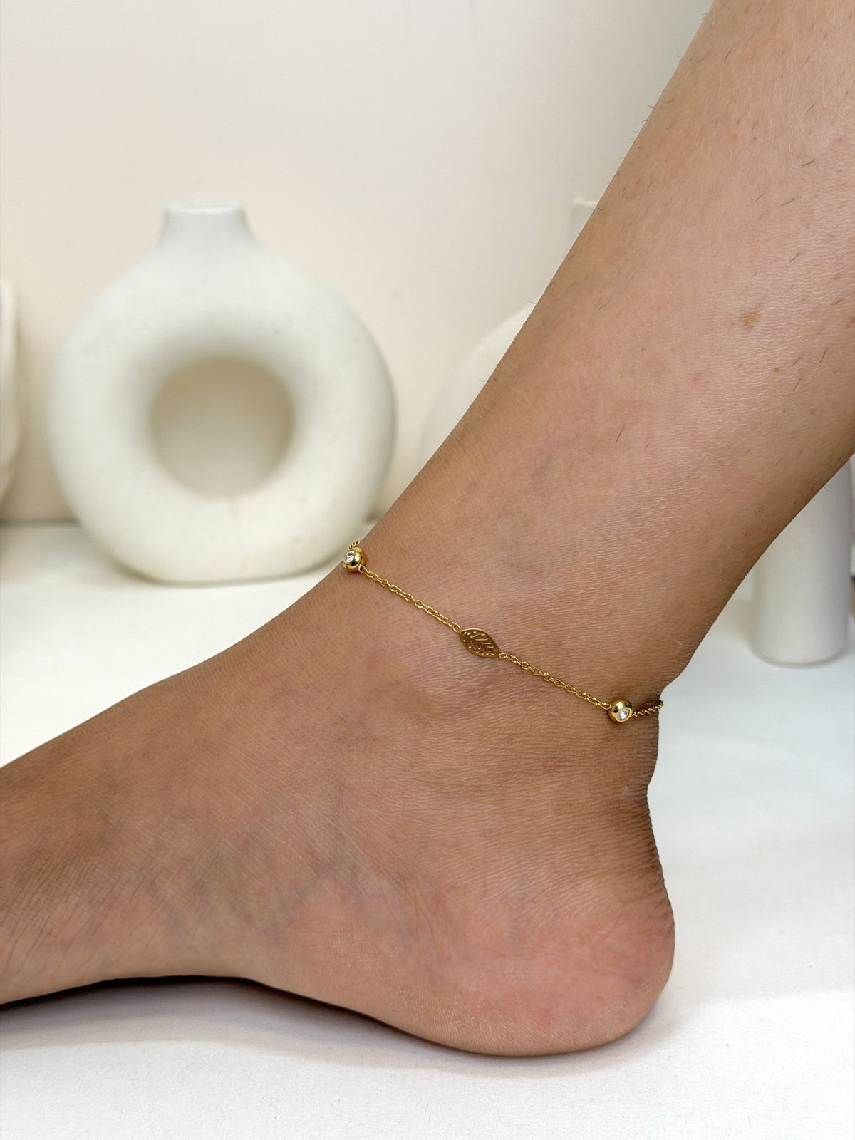 Paired Elvara Anklet in gold-tone, worn on the ankle with a fine chain, a leaf-shaped charm and two small beads