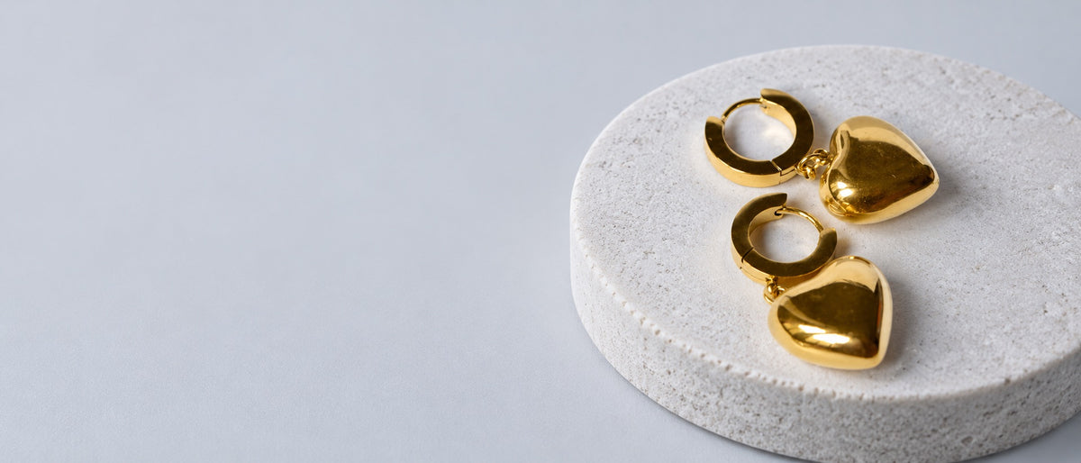 Paired Luxe Essentials gold heart huggie earrings on a pale stone plinth