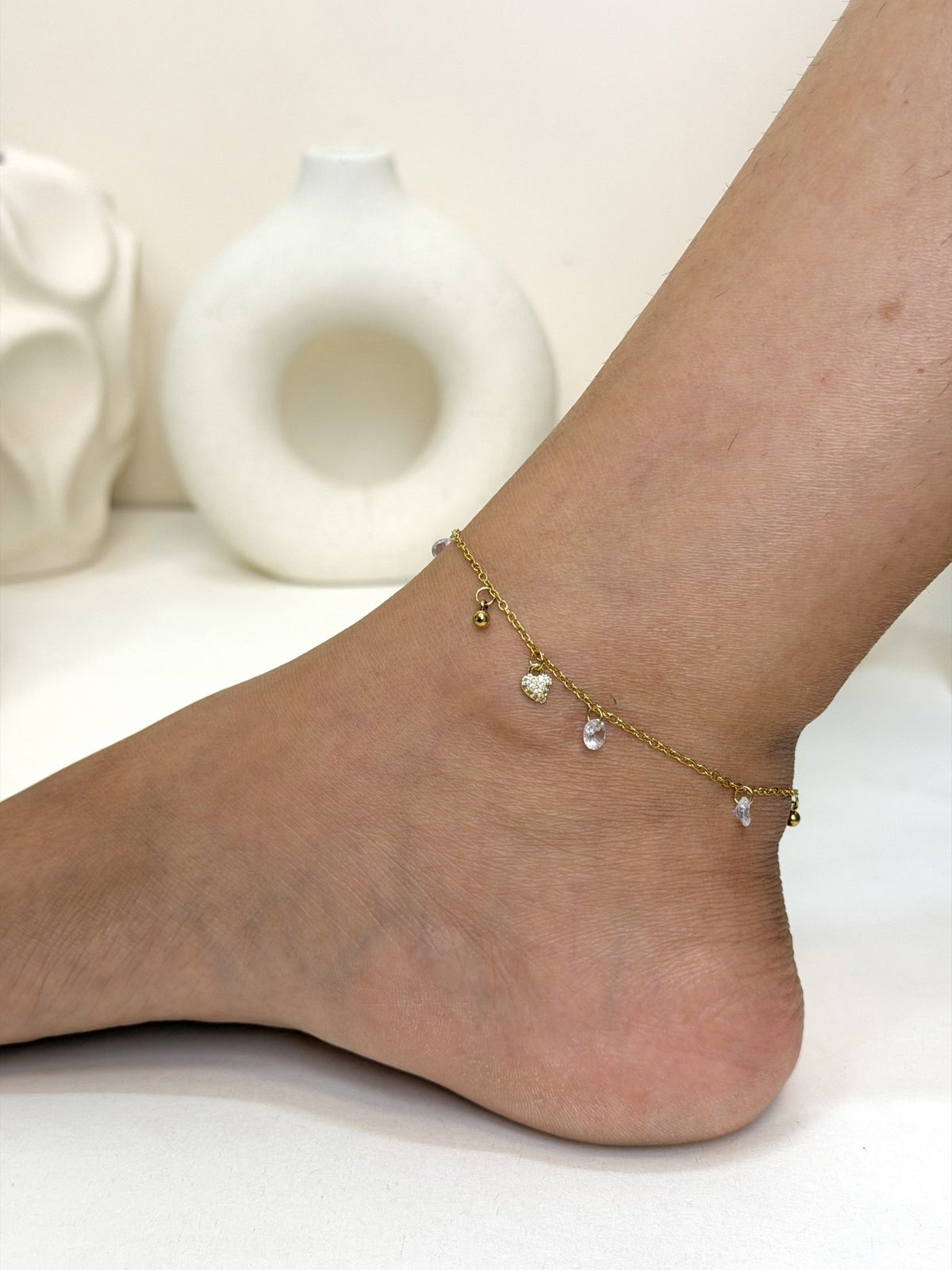 Paired Lunelle Anklet in gold-tone, worn on the ankle showing the heart charm and clear drop stones