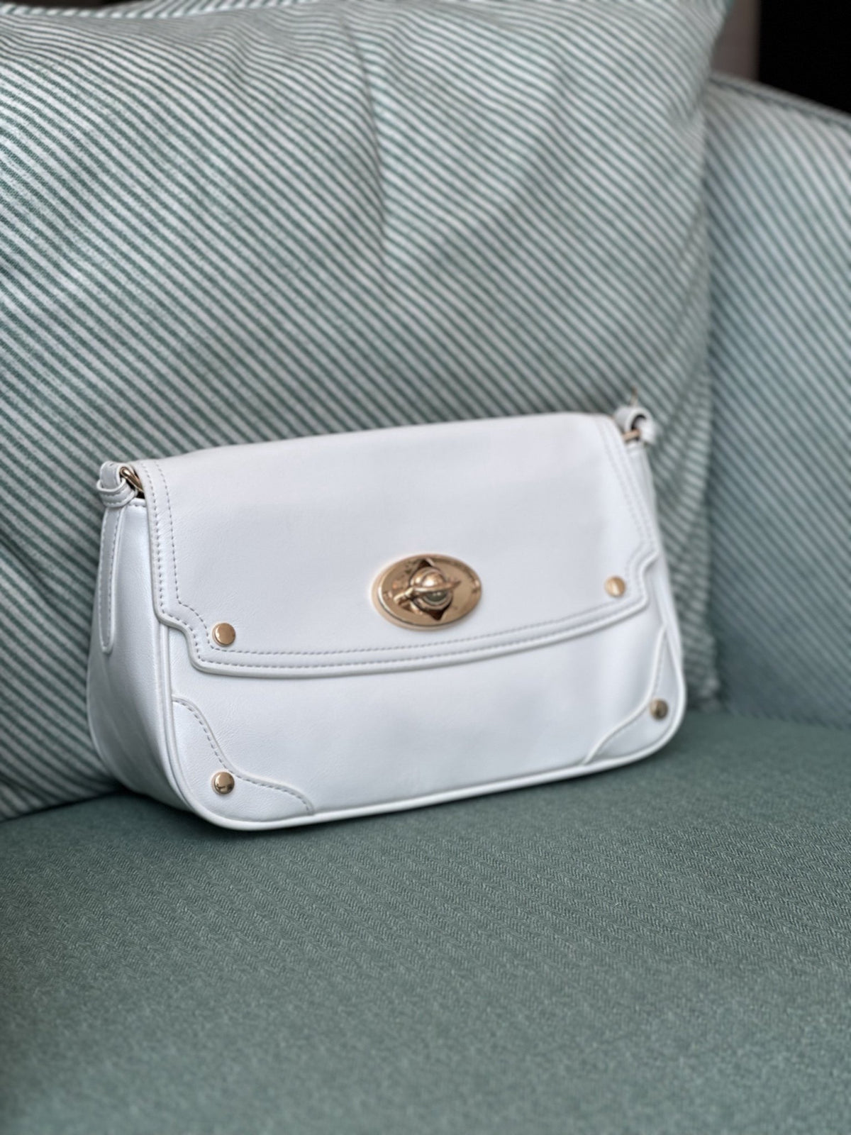 Paired Riviera Lock Bag in white, with a flap, gold-tone oval turn-lock and studded corners, resting on a striped armchair