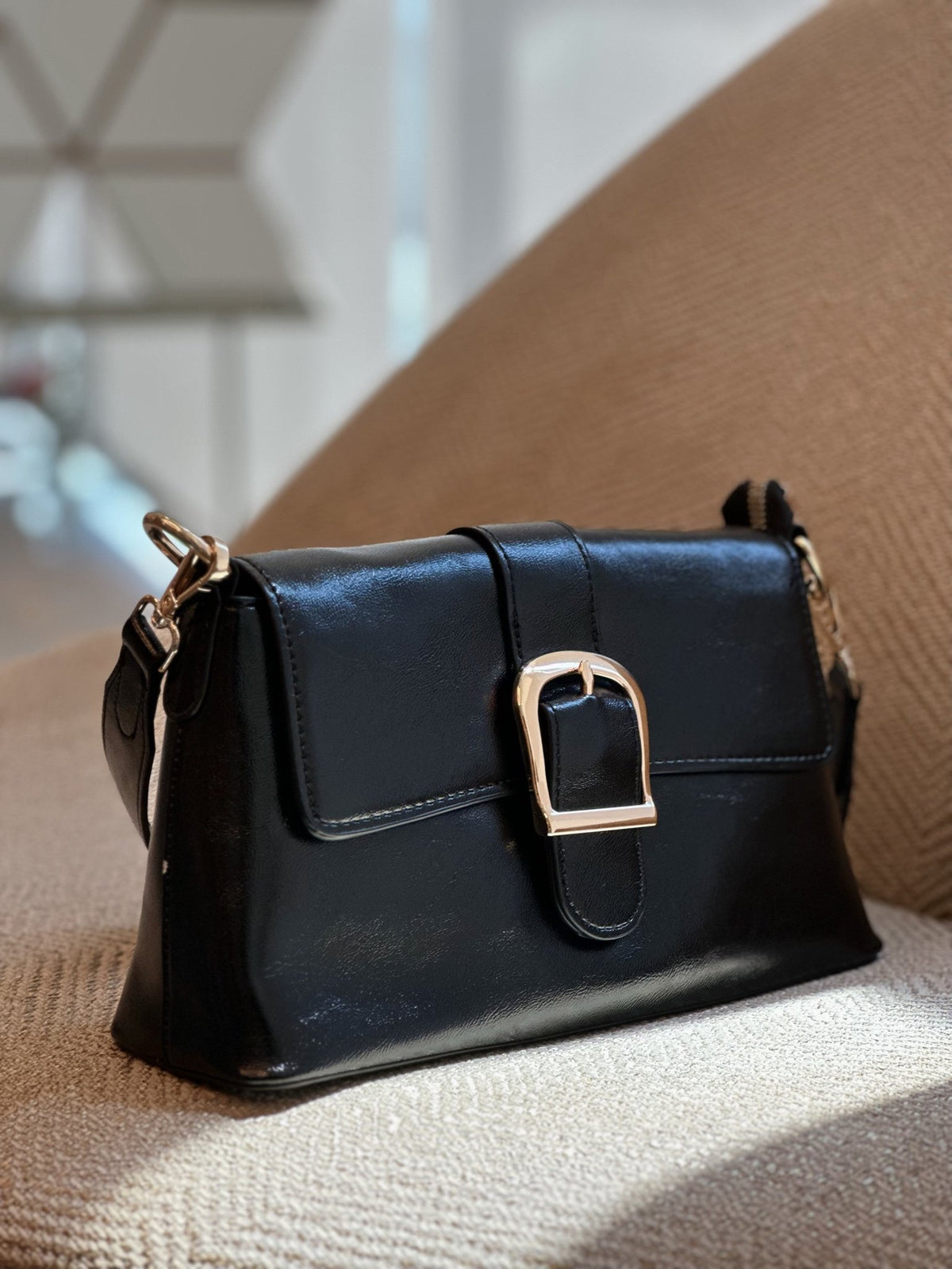 Paired Valora Buckle Sling Bag in black, with a buckled front flap and strap fittings, resting on a textured chair