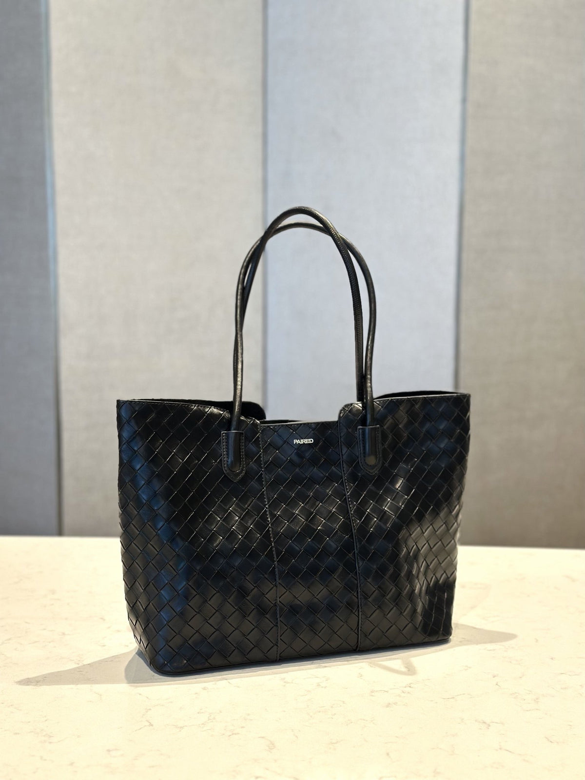 Paired Virelle Woven Bag in black, an open-top tote with two rolled handles and a woven texture, standing on a marble table