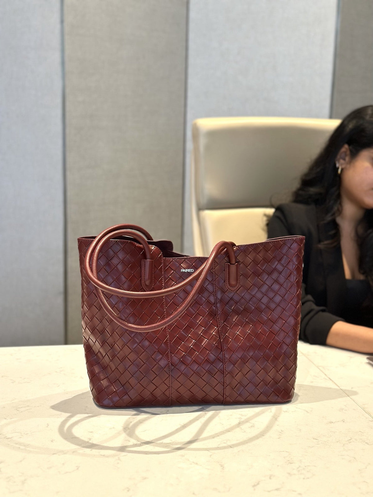 Paired Virelle Woven Bag in maroon, an open-top tote with two rolled handles and a woven texture, seen on a marble table