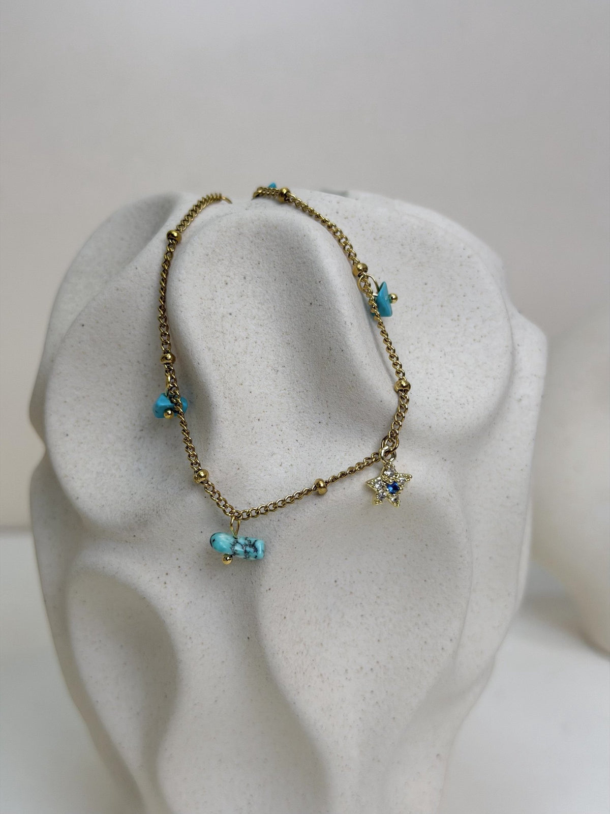 Paired Alvara Anklet in gold-tone, close view on a textured vase with turquoise-coloured stones and a star charm