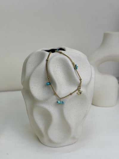 Paired Alvara Anklet in gold-tone, draped over a textured cream vase showing turquoise charms and a star