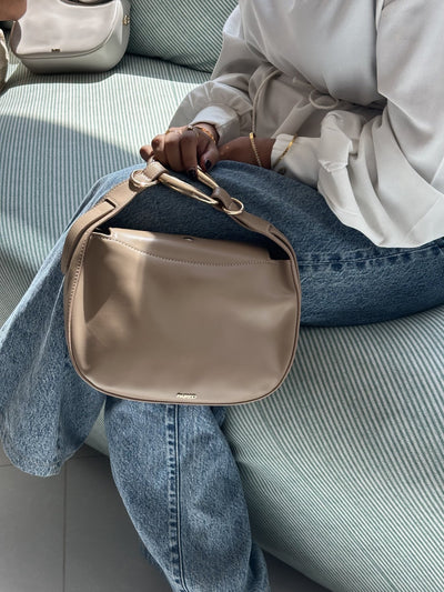 Paired Arden Soft Bag in taupe apricot, held by the ring handle while seated, styled with a white shirt and jeans
