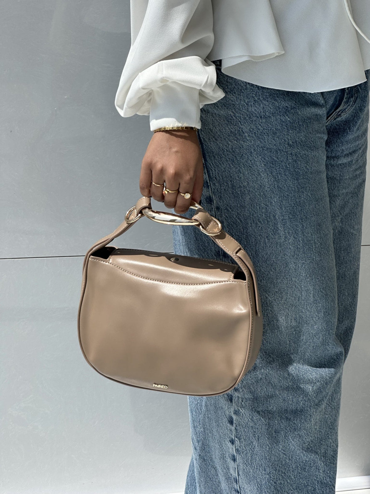 Paired Arden Soft Bag in taupe apricot, held by the ring handle against jeans and a white sleeve, photographed sideways