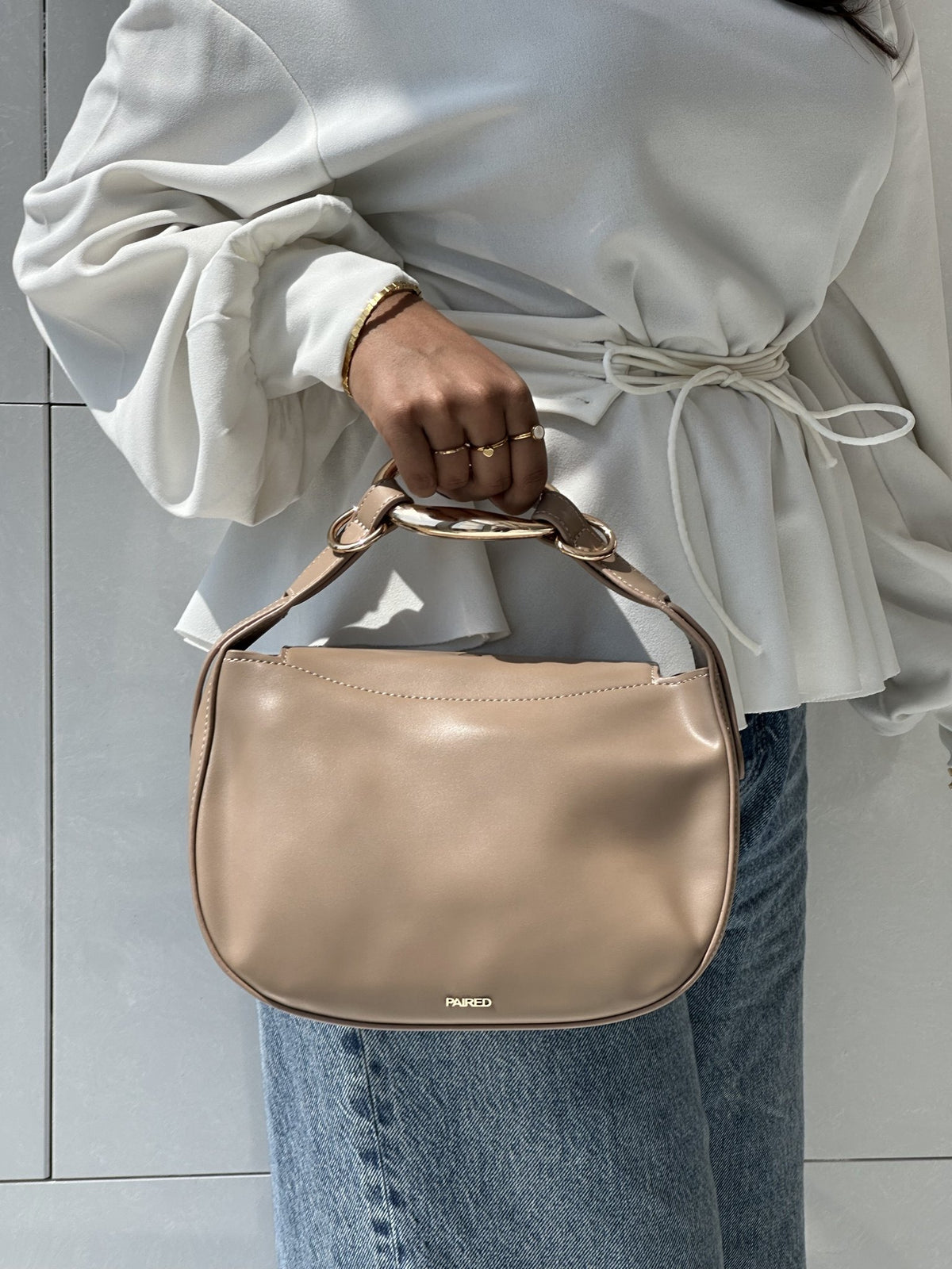 Paired Arden Soft Bag in taupe apricot, held by the ring handle against jeans and a white blouse, photographed sideways