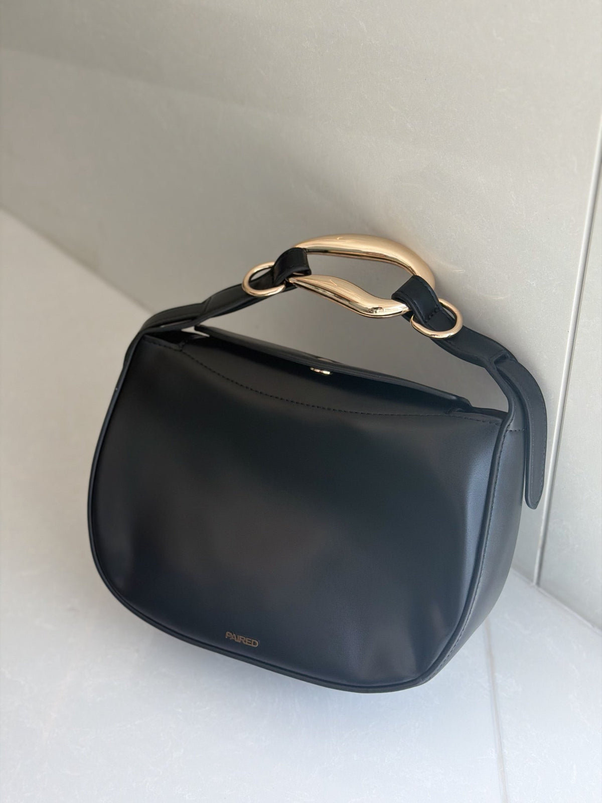 Paired Arden Soft Bag in black, with a soft half-moon shape and a sculptural gold-tone ring handle, on a white surface