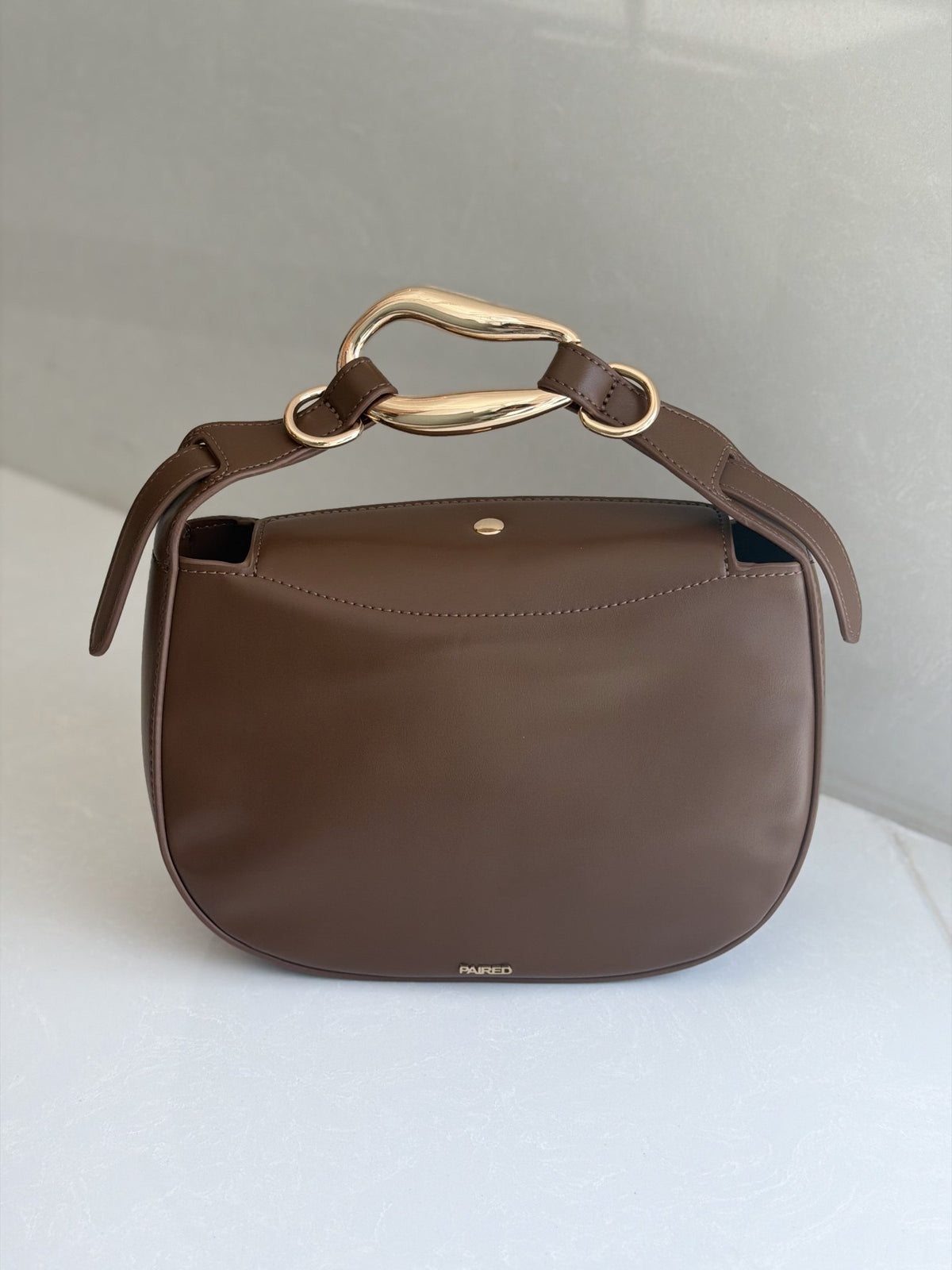 Paired Arden Soft Bag in coffee brown, with a soft rounded shape and a sculptural gold-tone ring handle, shown from the front