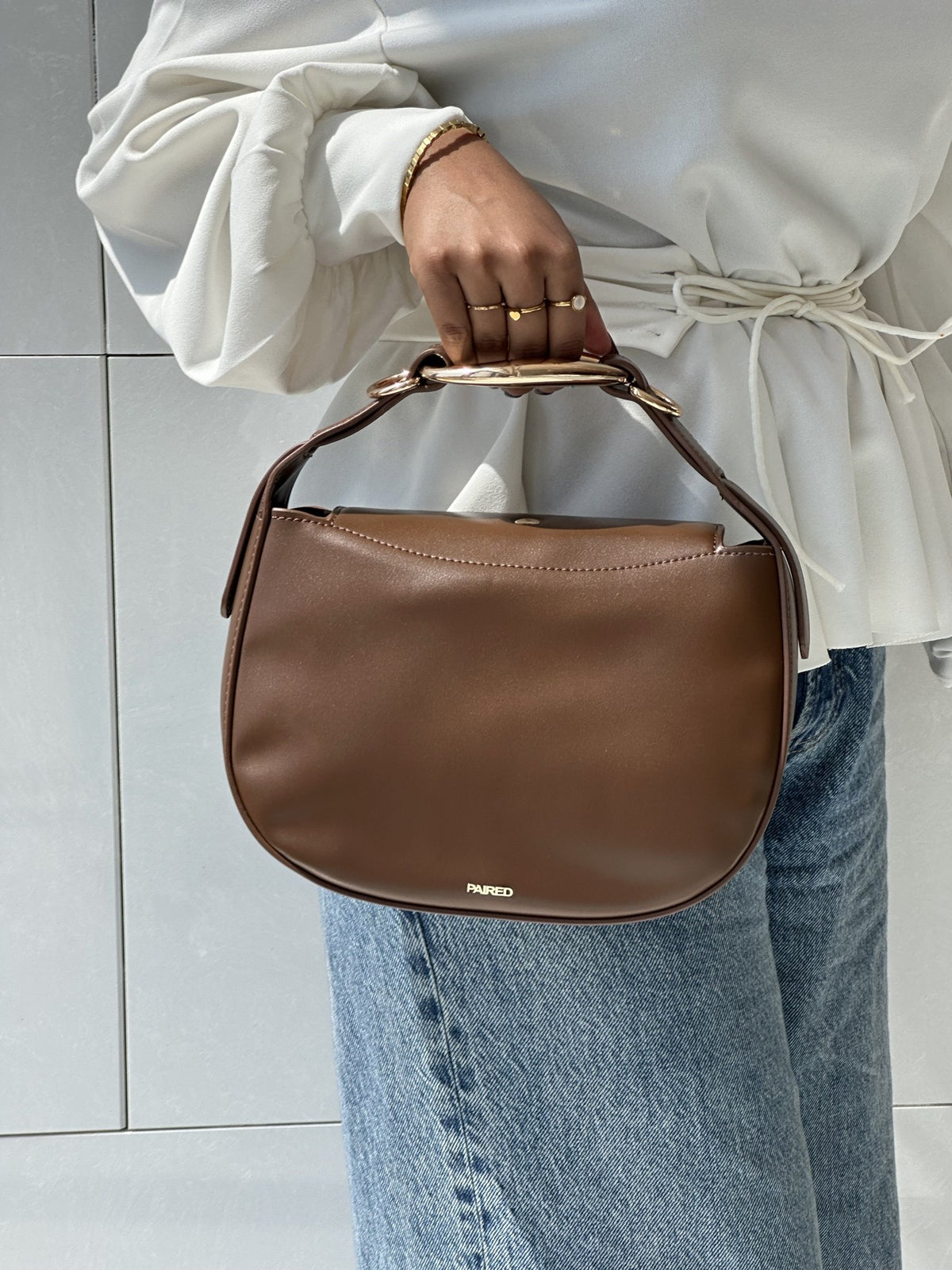 Paired Arden Soft Bag in coffee brown, held by the ring handle against jeans and a white blouse, photographed sideways