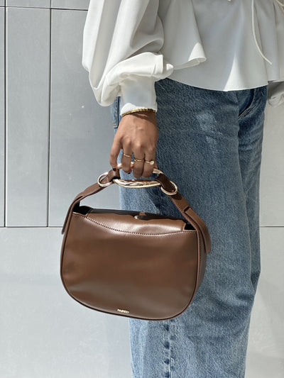 Paired Arden Soft Bag in coffee brown, held by the ring handle against jeans and a white blouse, photographed sideways