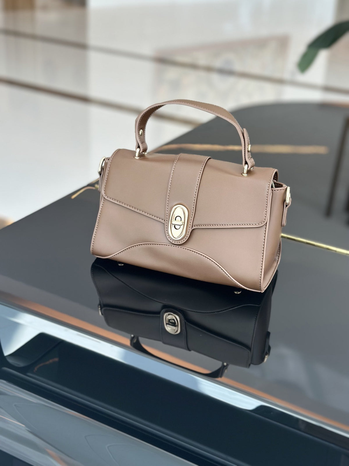 Paired Aurea Crest Bag in taupe apricot, with a top handle and gold-tone oval turn-lock, on a glossy black surface