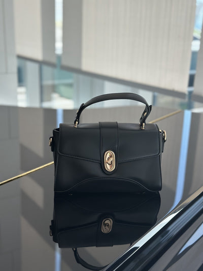 Paired Aurea Crest Bag in black, with a top handle and gold-tone oval turn-lock on a glossy dark surface