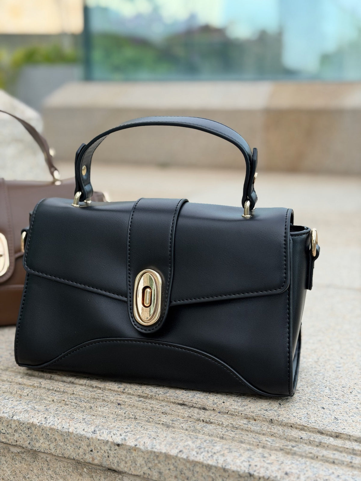 Paired Aurea Crest Bag in black, with a top handle and gold-tone oval turn-lock, set on a stone ledge