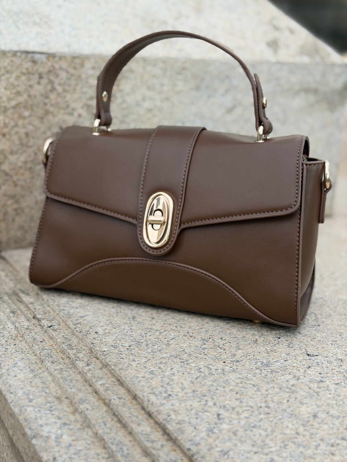 Paired Aurea Crest Bag in coffee brown, with a top handle, curved flap and gold-tone oval turn-lock, resting on a stone ledge
