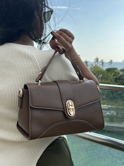 Paired Aurea Crest Bag in coffee brown, worn on the shoulder with a top handle and gold-tone turn-lock, photographed sideways
