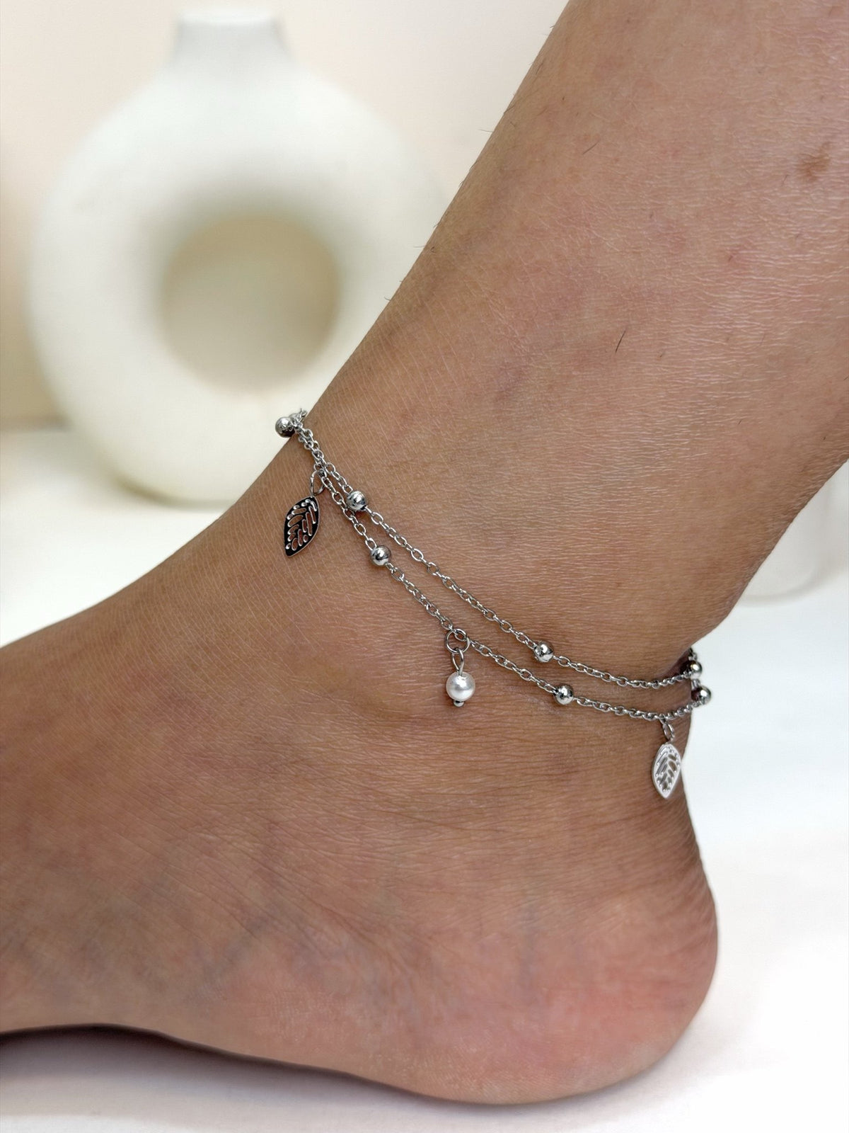 Paired Aurevia Anklet in silver-tone, worn on the ankle with layered beaded chains, a leaf charm and small drops