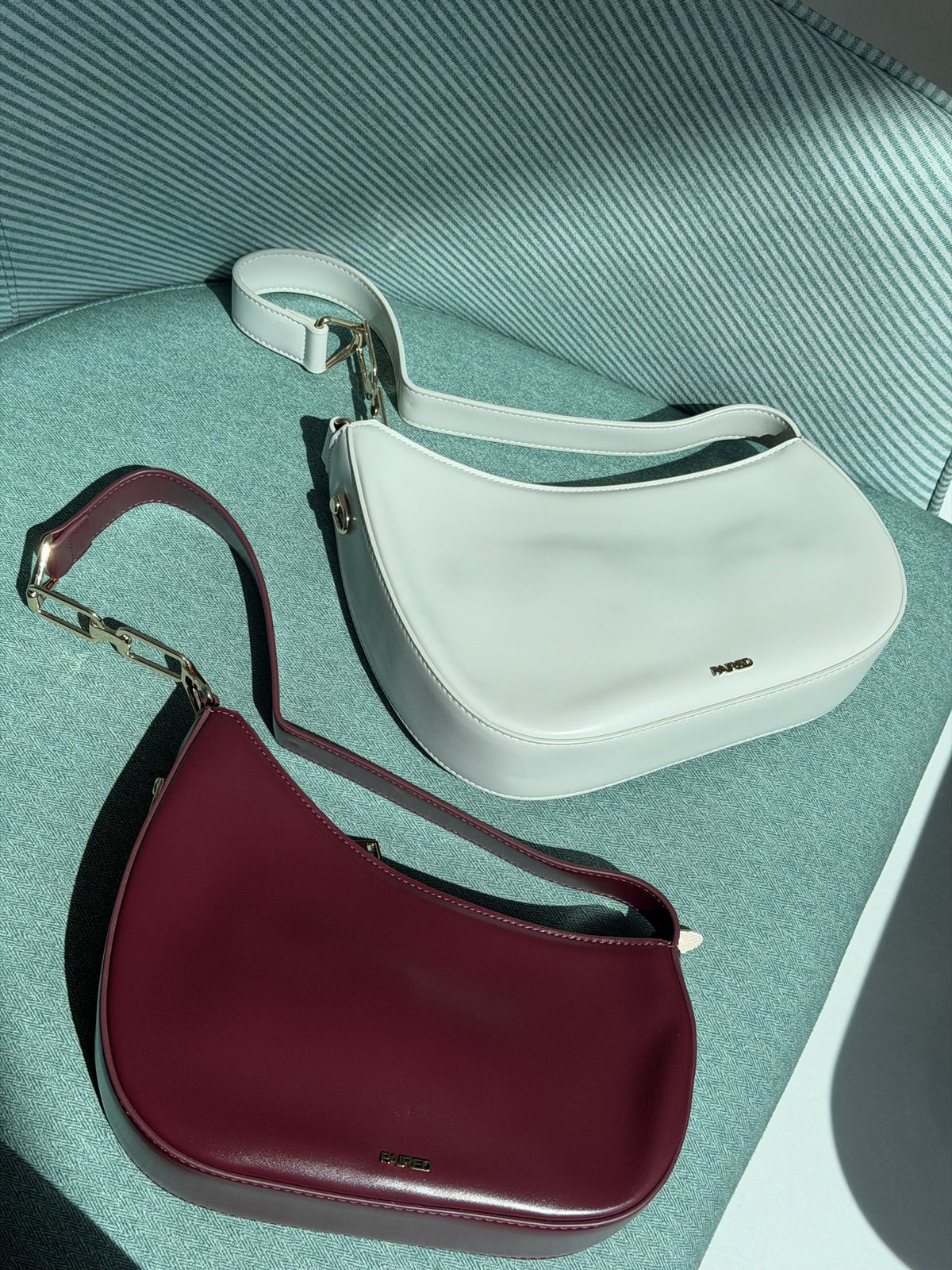 Paired Clarissa Chain Bag in white and burgundy lying together on a teal armchair, each with a flat strap and chain link