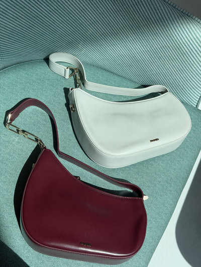 Paired Clarissa Chain Bag in white and burgundy lying together on a teal armchair, each with a flat strap and chain link