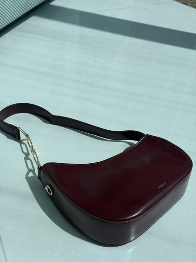 Paired Clarissa Chain Bag in burgundy, a crescent shoulder bag with a flat strap and chunky chain link on a pale surface