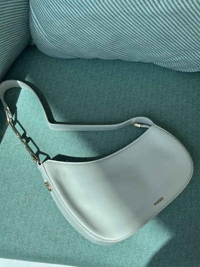 Paired Clarissa Chain Bag in white, a crescent shoulder bag with a flat strap and chunky chain link, lying on a teal armchair