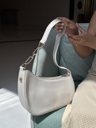 Paired Clarissa Chain Bag in white, held by the flat strap and gold-tone chain link on a teal chair, photographed sideways