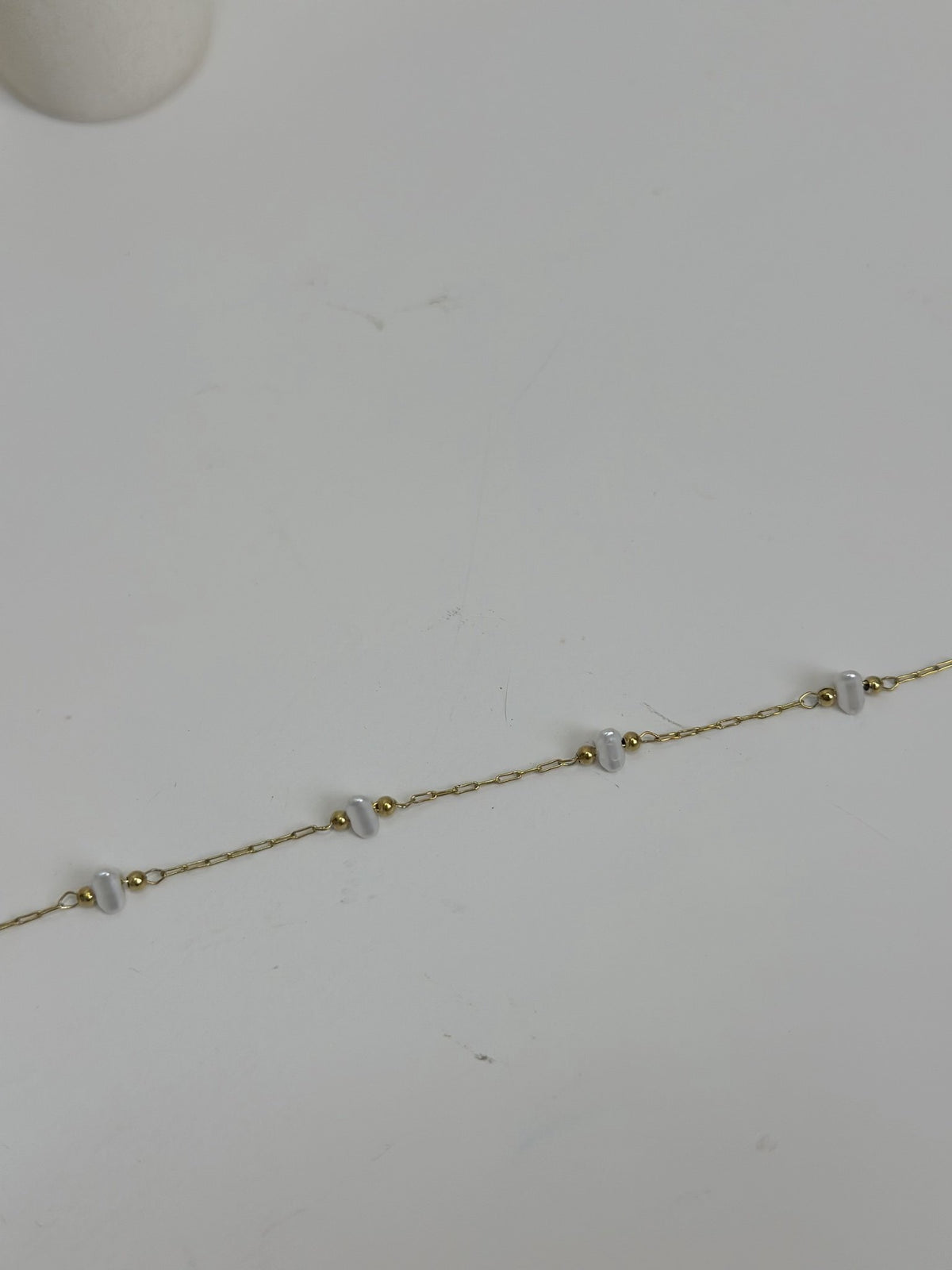 Paired Dorevia Bracelet in gold-tone, laid flat on a grey surface showing a row of white heart beads on a slim chain