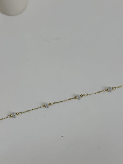 Paired Dorevia Bracelet in gold-tone, laid flat on a grey surface showing a row of white heart beads on a slim chain