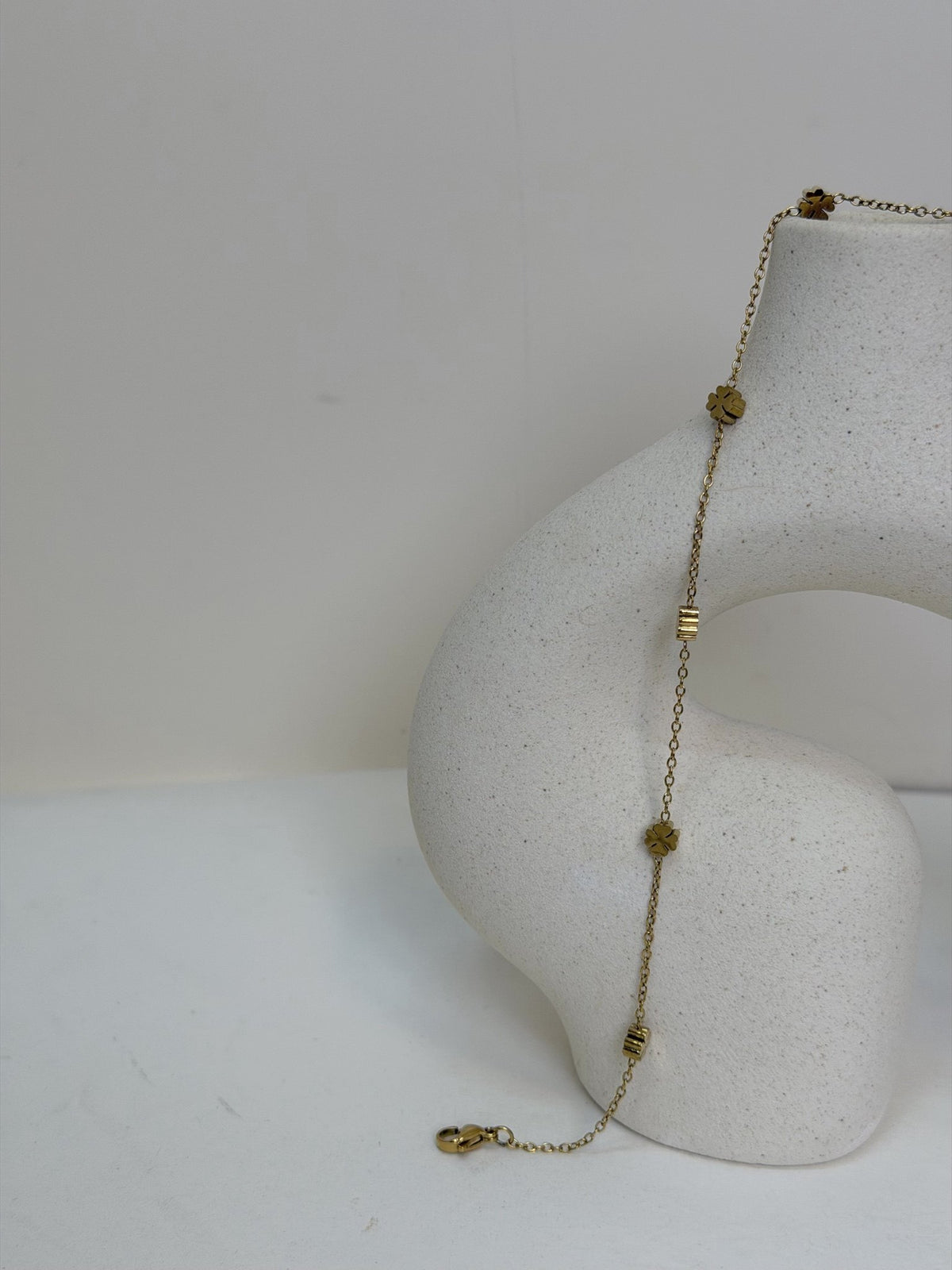 Paired Elara Anklet in gold-tone, draped over a cream sculptural vase showing flower charms and a lobster clasp