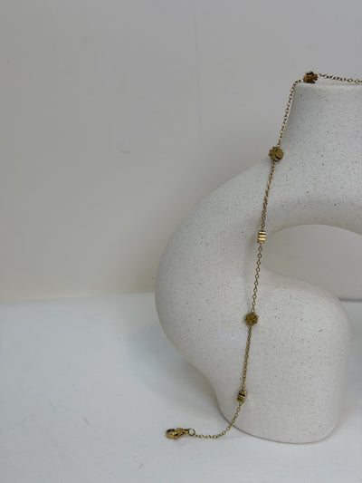 Paired Elara Anklet in gold-tone, draped over a cream sculptural vase showing flower charms and a lobster clasp