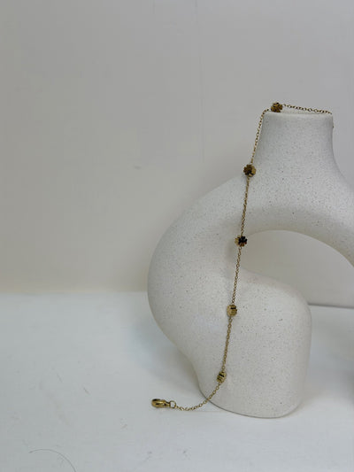 Paired Elara Anklet in gold-tone, draped on a cream sculptural vase showing flower charms and the lobster clasp
