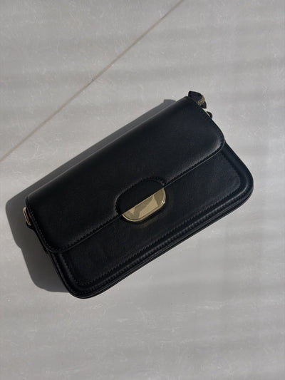 Paired Elise Lock Bag in black, laid flat on a pale surface, showing the smooth flap, side buckle and strap