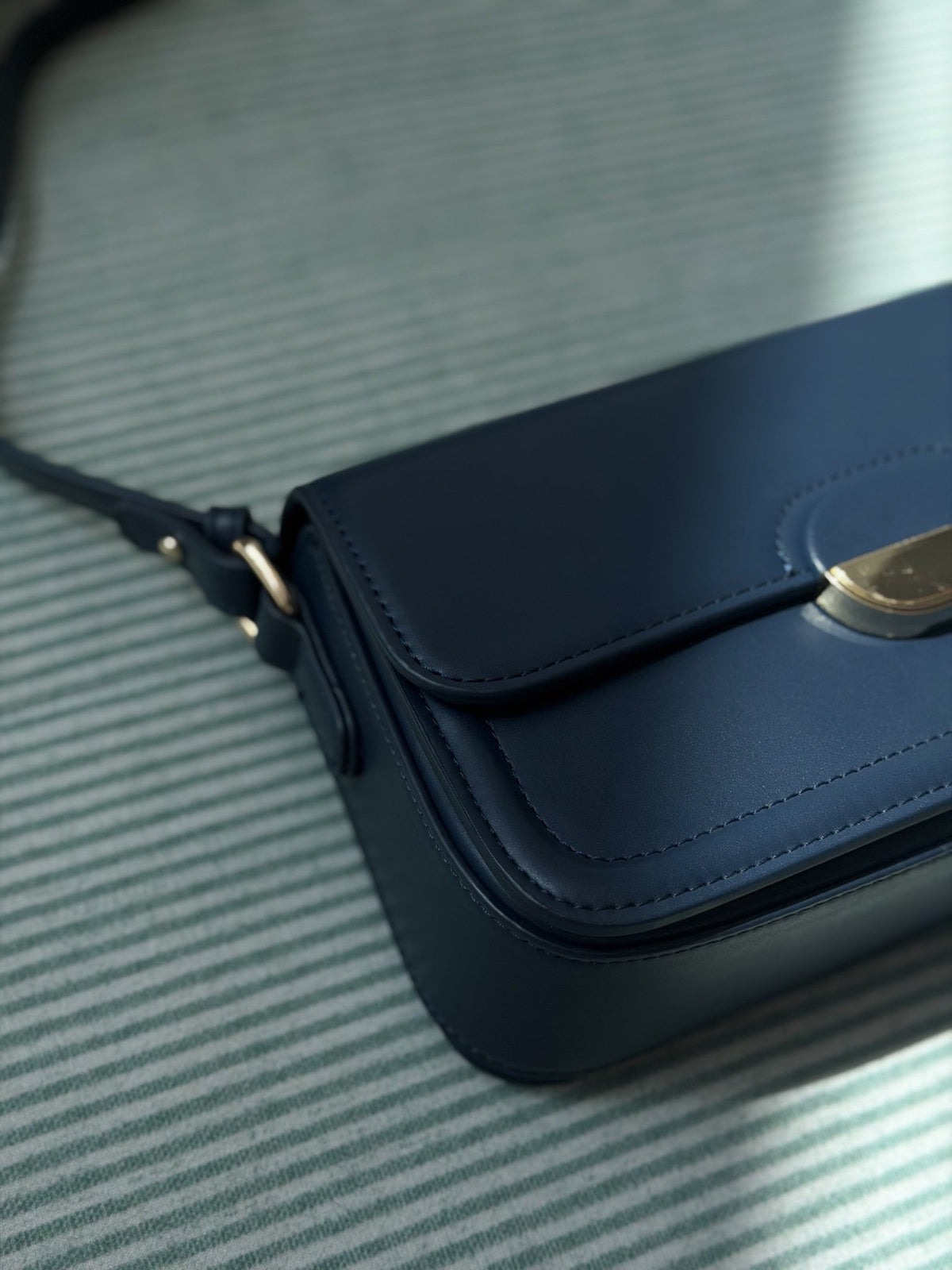 Paired Elise Lock Bag in navy blue, close-up of the flap and gold-tone half-moon lock plate on a striped cloth