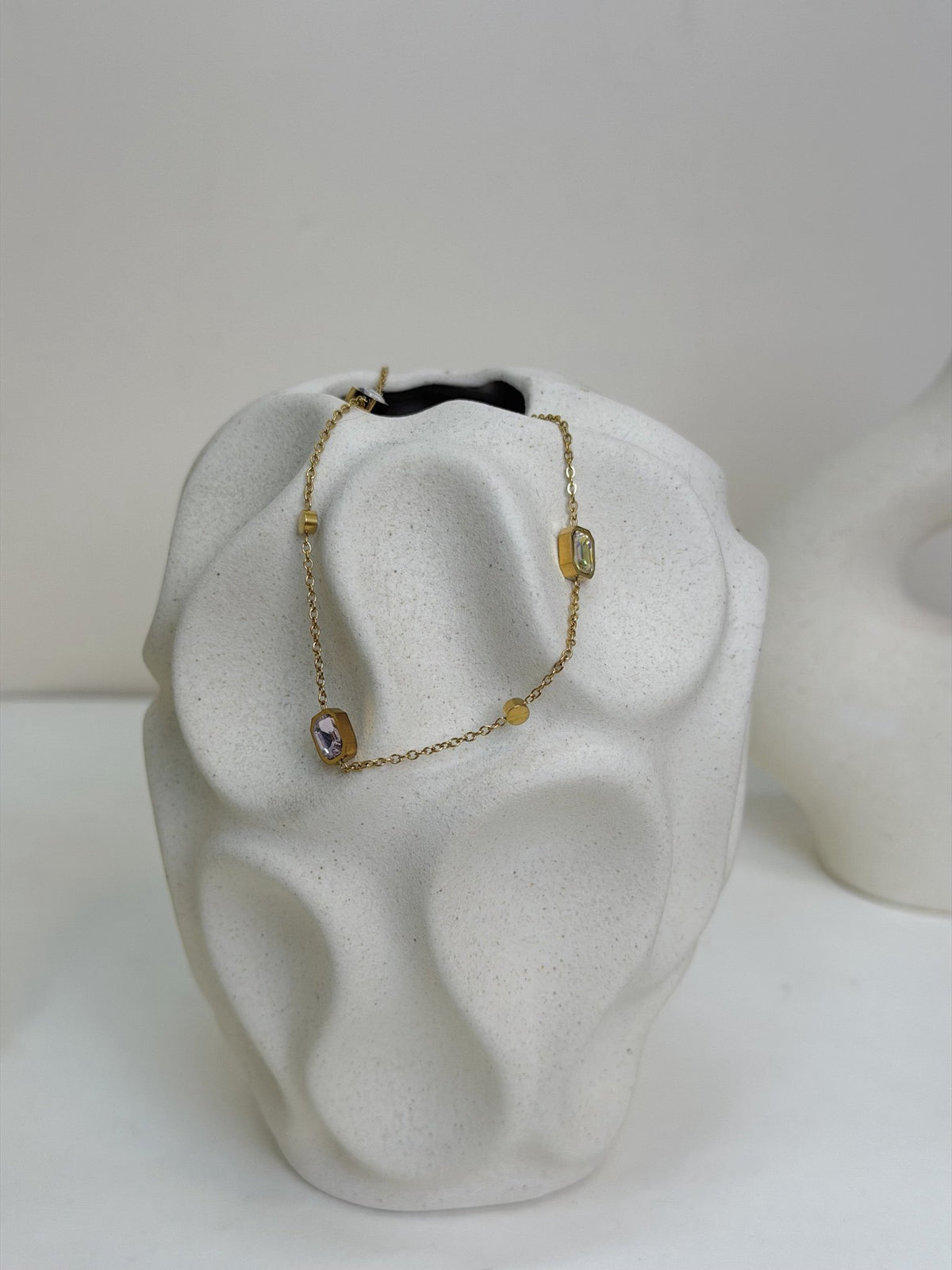 Paired Evania Anklet in gold-tone, draped over a textured cream vase showing pink and clear stones