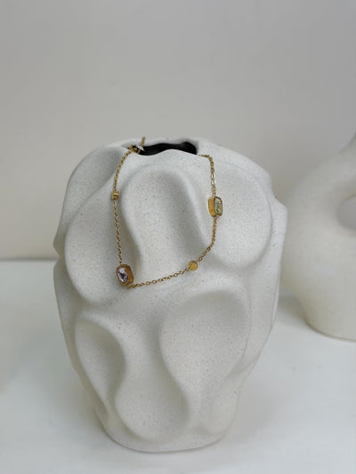 Paired Evania Anklet in gold-tone, draped over a textured cream vase showing pink and clear stones