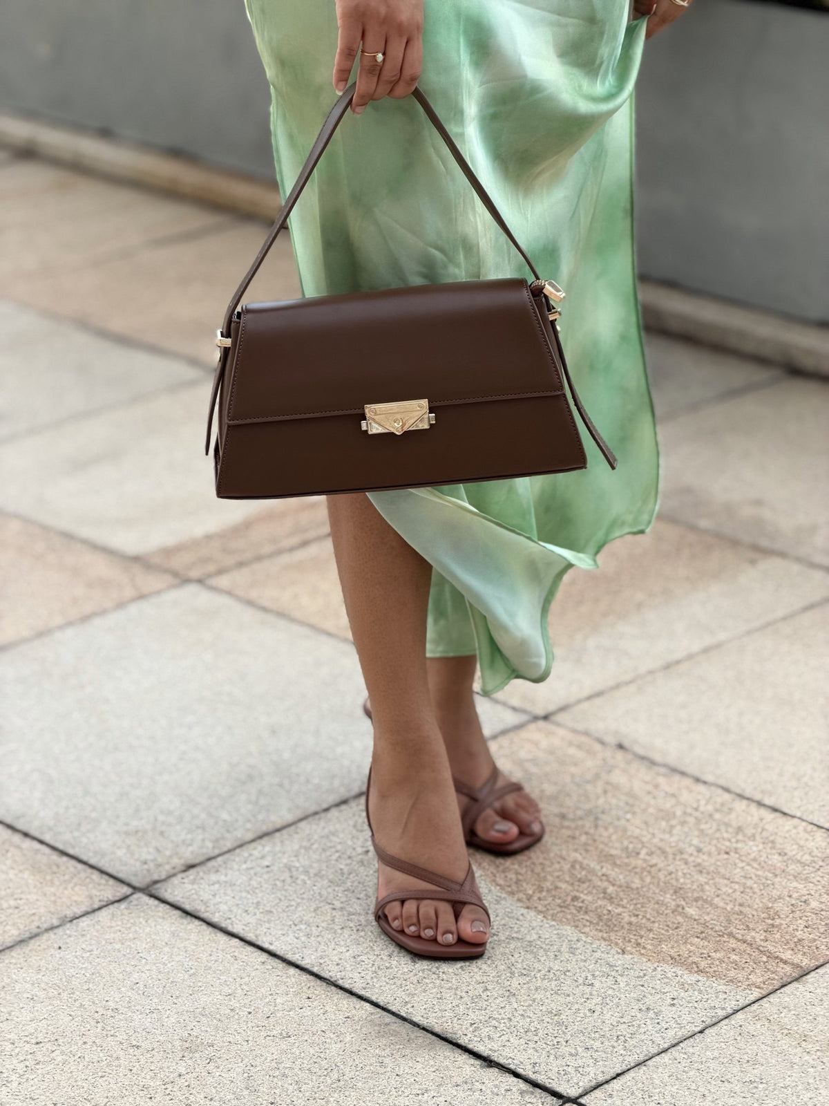 Paired Liora Frame Bag in coffee brown, carried by a slim strap in the hand beside a green satin skirt