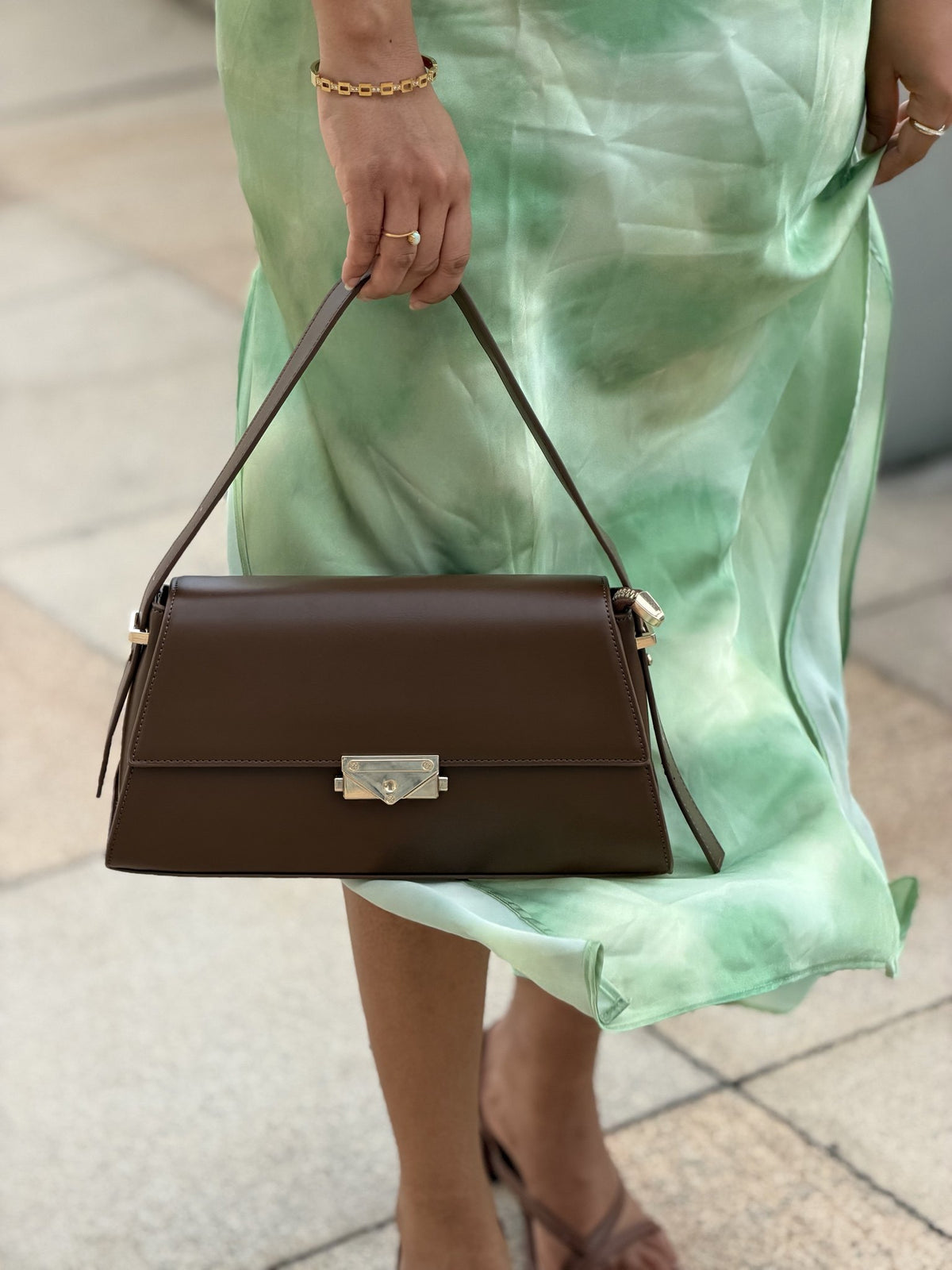Paired Liora Frame Bag in coffee brown, held by the strap at the front, showing the angled flap and gold-tone clasp