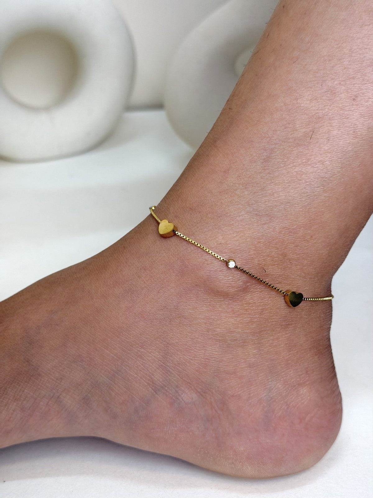 Paired Lorielle Anklet in gold-tone, worn on the ankle with a fine chain, small heart charms and a clear stone