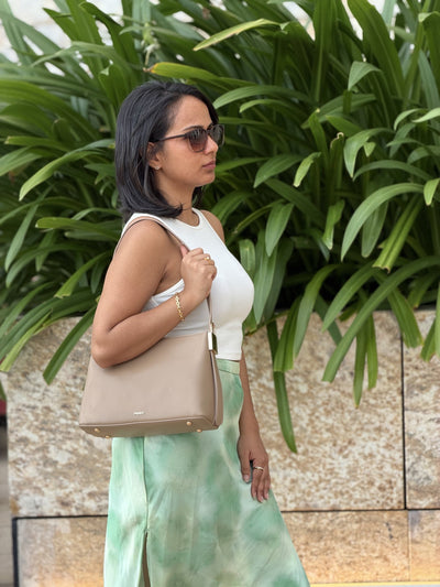 Paired Oriana Gold-Lock Bag in taupe apricot, worn on the shoulder, side view, with a white tank top and plants behind