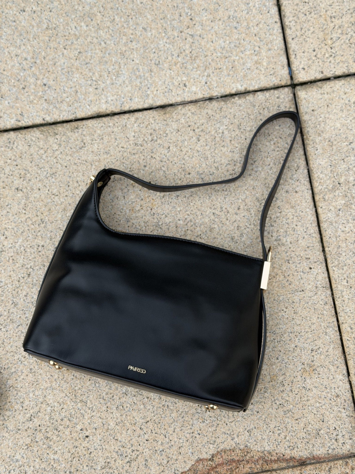 Paired Oriana Gold-Lock Bag in black, a shoulder bag lying on stone paving with its strap and small gold-tone base detail