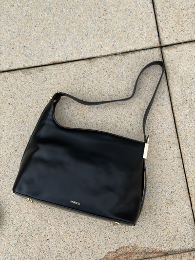 Paired Oriana Gold-Lock Bag in black, a shoulder bag lying on stone paving with its strap and small gold-tone base detail